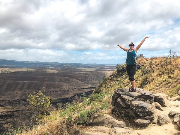 Brittany on a rock with arms raised excited about the views of mountains and valleys behind her on the way to the hike to Waipo'o Falls in Kauai Hawaii